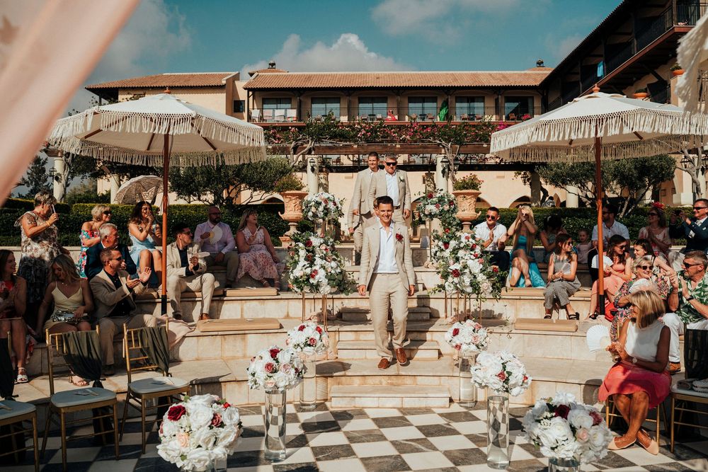 Outdoor wedding ceremony at a Mediterranean venue with checkered floor and white umbrellas surrounded by guests.