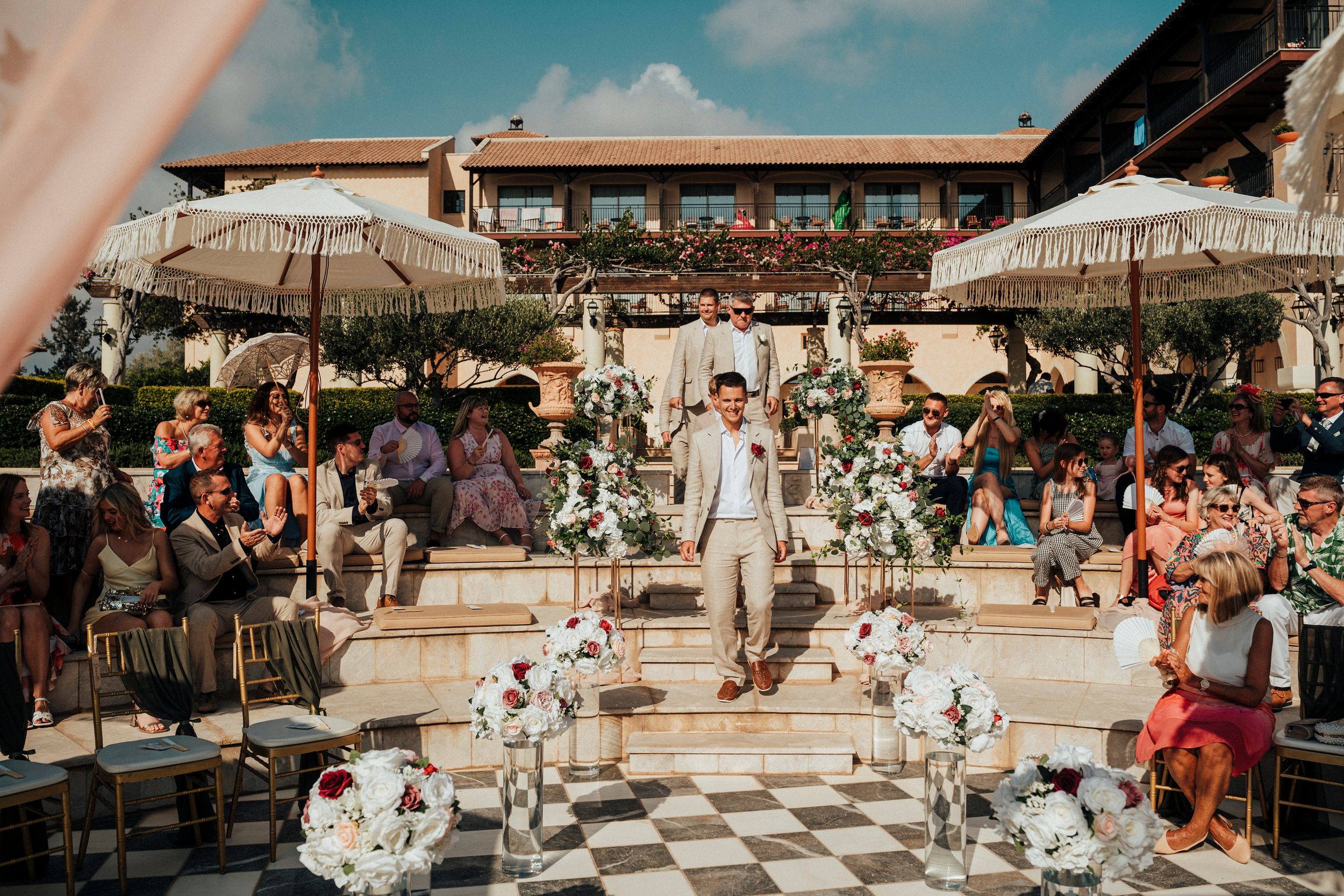 Outdoor wedding ceremony at a Mediterranean venue with checkered floor and white umbrellas surrounded by guests.