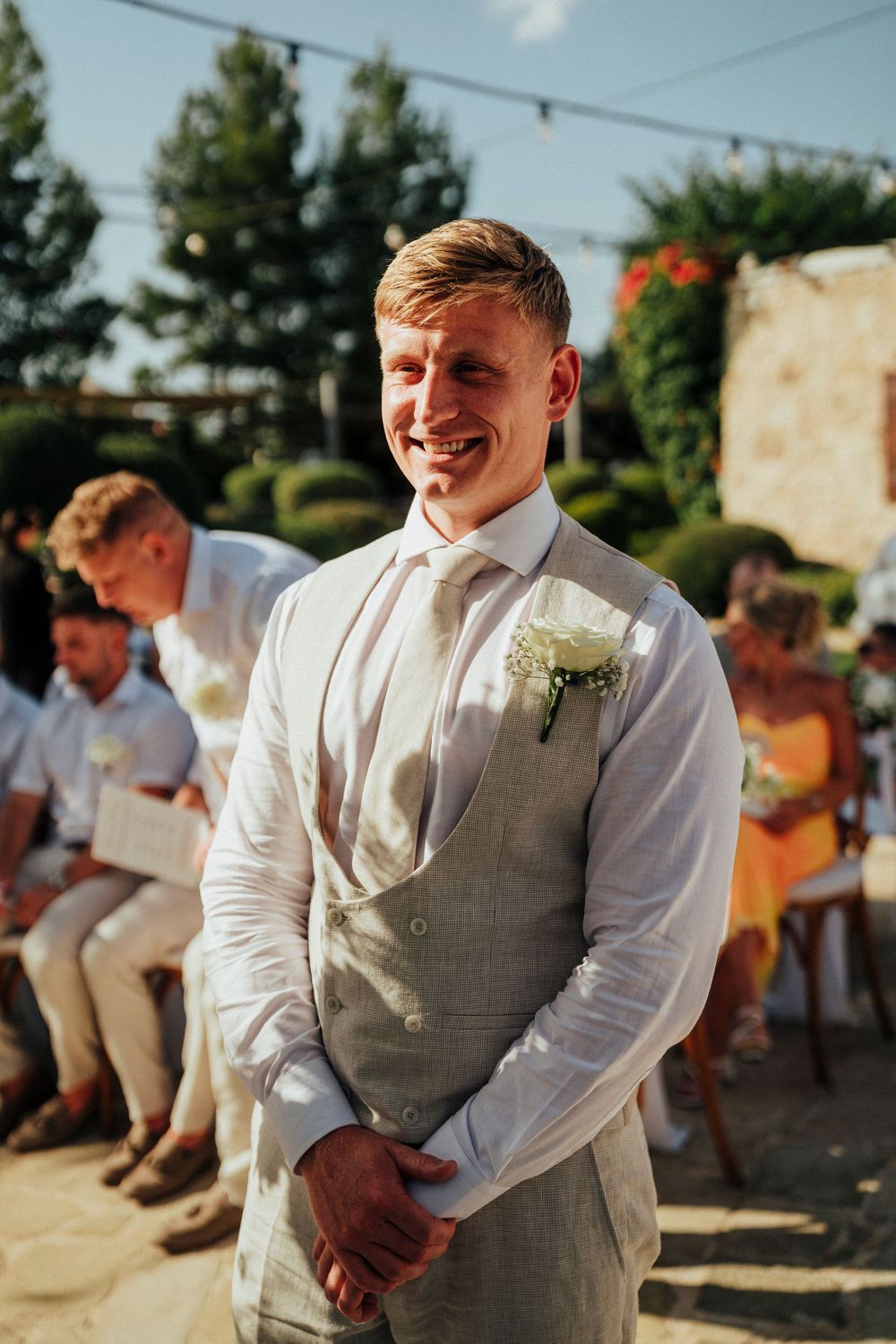 A groomsman in a white shirt and gray vest smiles joyfully at an outdoor wedding ceremony.