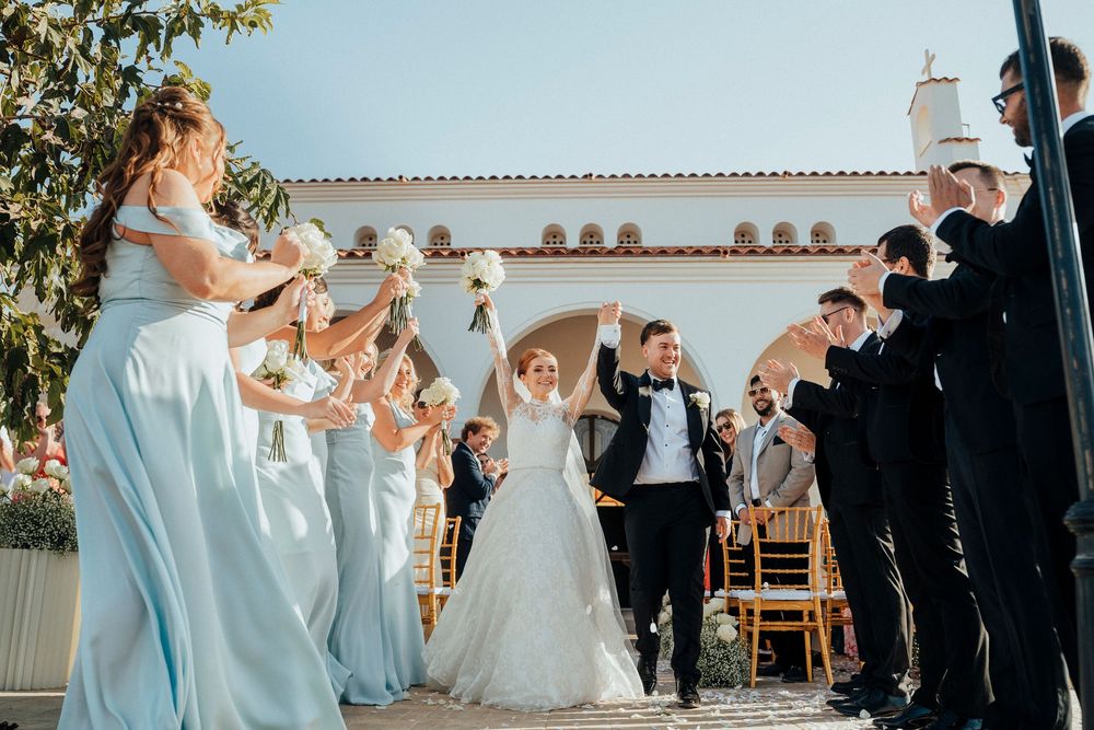 Wedding party poses together in formal attire outdoors on a sunny day with white walls and greenery in background.