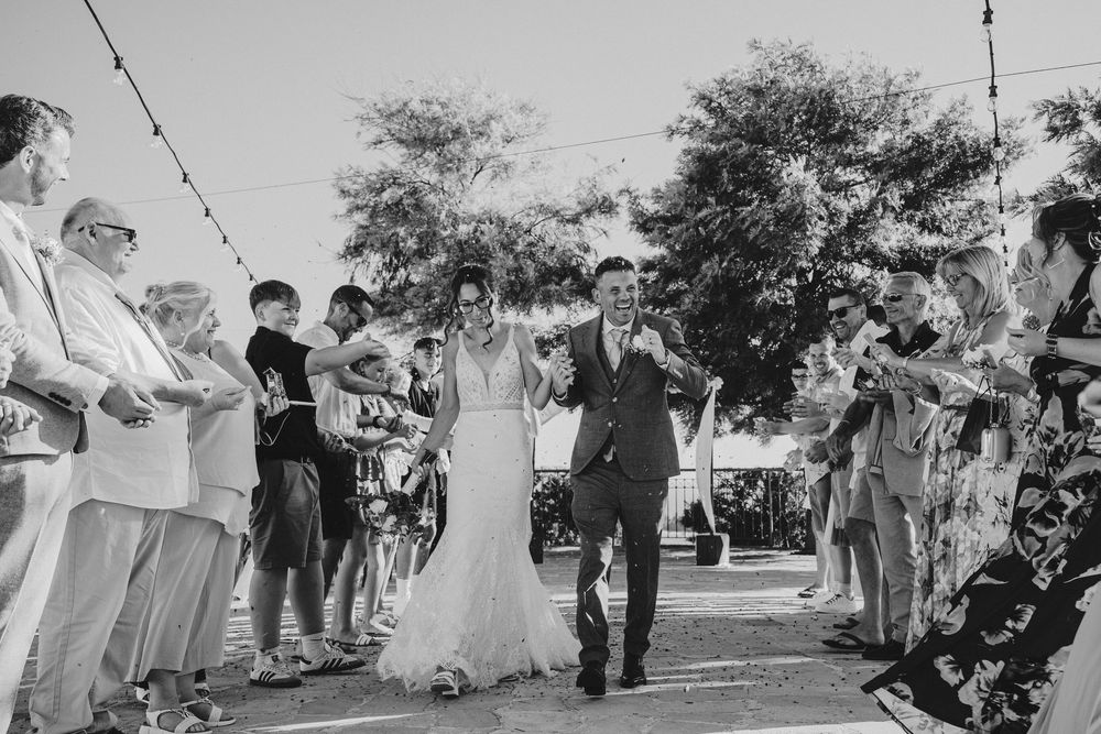 A black and white wedding ceremony photo showing guests seated outdoors under trees during a daytime celebration.