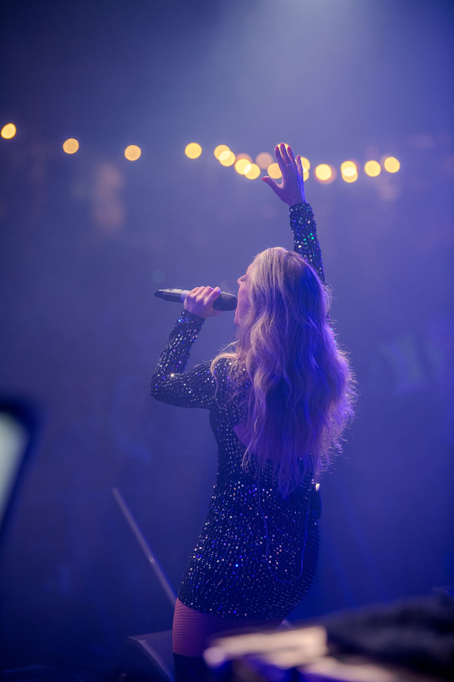 Performer in sparkly black outfit on stage illuminated by purple lighting and bokeh effects during concert performance.