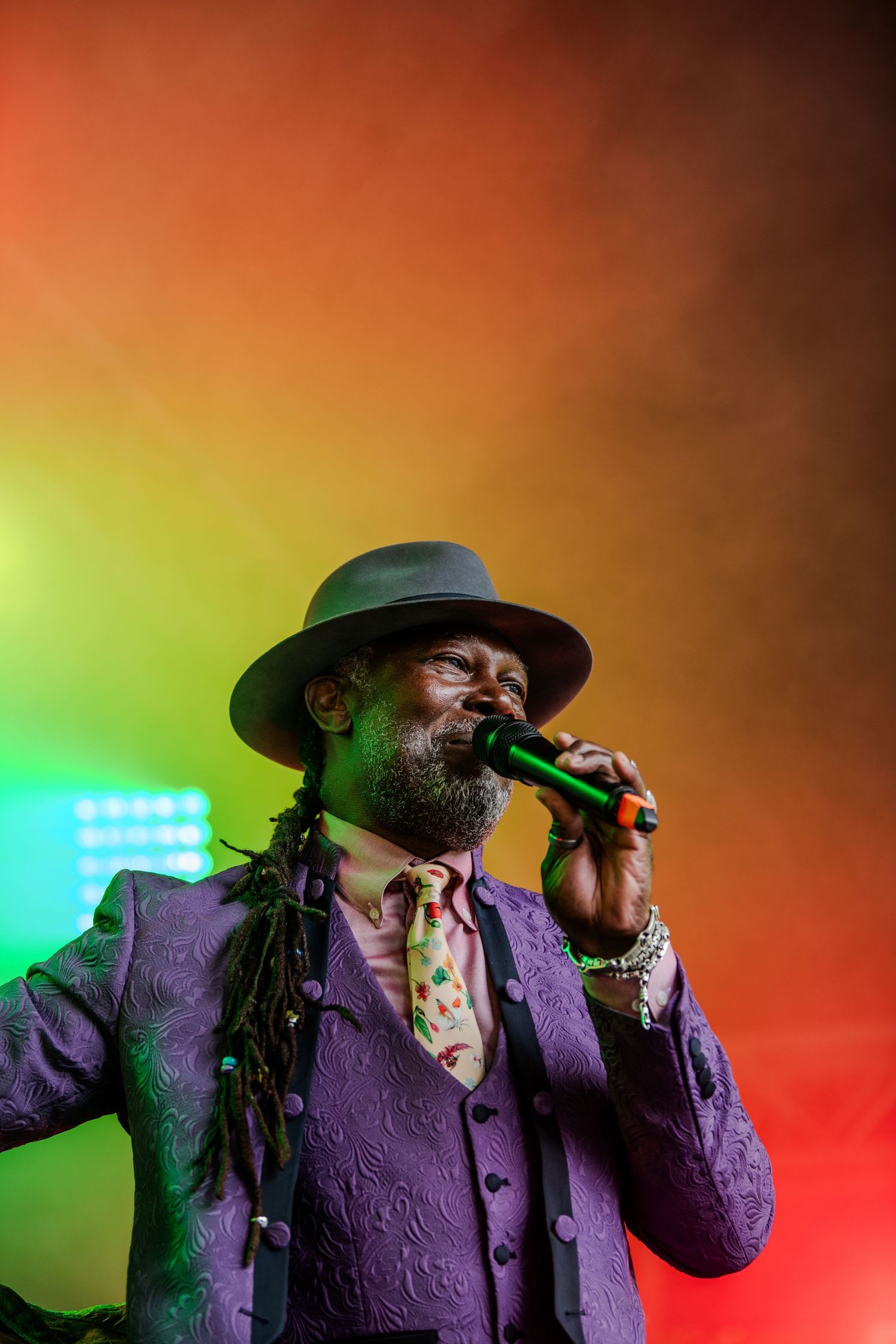 Performer in a purple jacket and hat singing passionately on stage with colorful lighting effects.