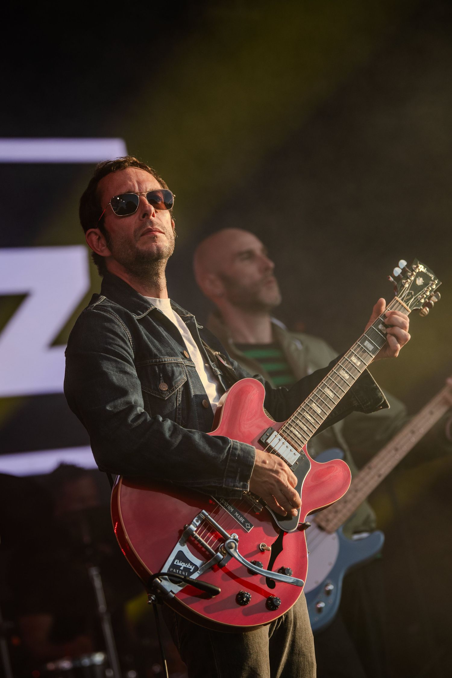 Guitarist in sunglasses plays red electric guitar during performance on dimly lit stage.