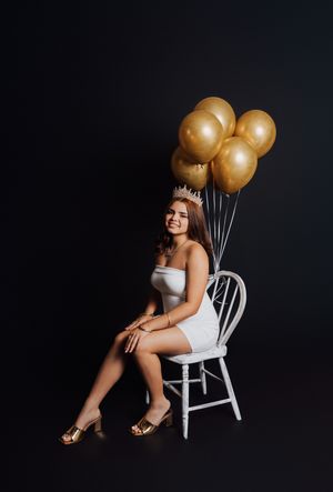 Young woman in white dress and gold crown sits on white chair holding gold balloons against black background.