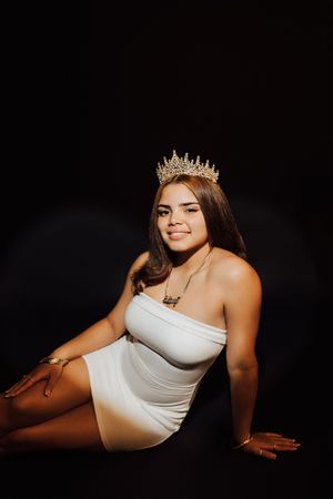 Young woman in white dress and gold crown poses confidently against black background.
