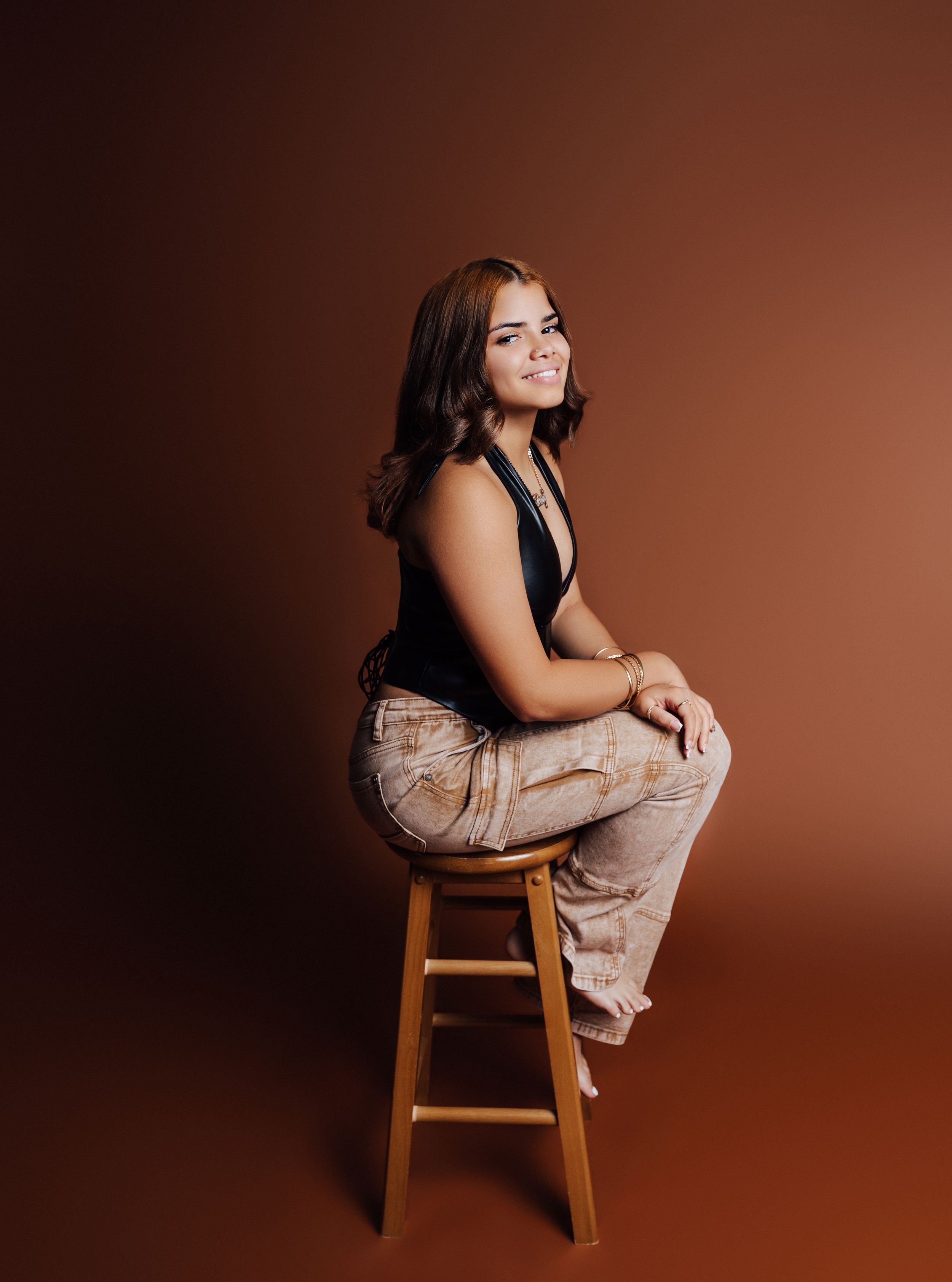 Young woman in black top and patterned pants sitting on wooden stool against warm brown studio background.