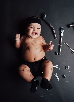 Smiling baby with grease marks lies on dark floor surrounded by mechanic tools, wearing black shorts and cap.