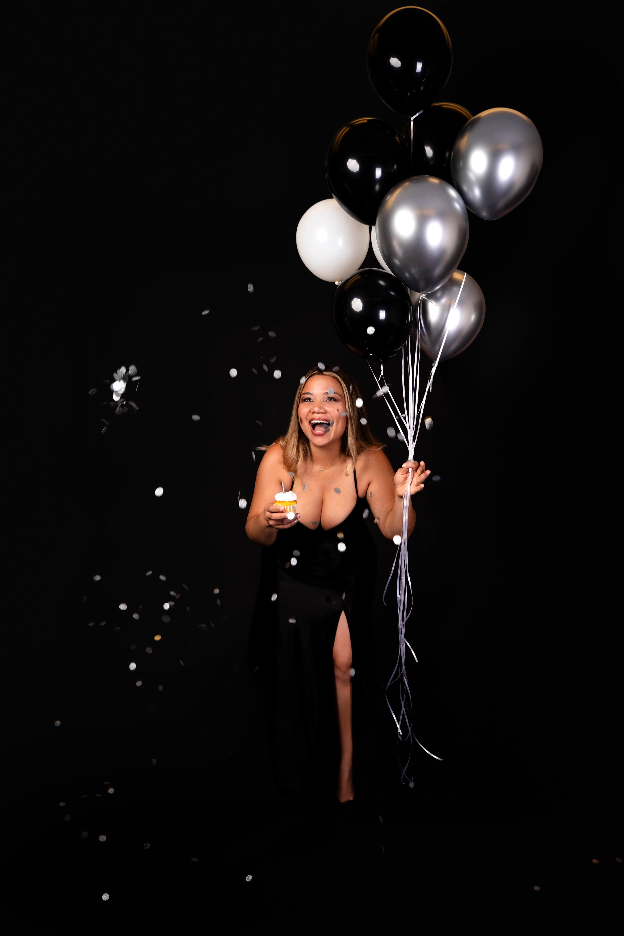 Joyful woman in black gown holding balloons and champagne with confetti on dark background.