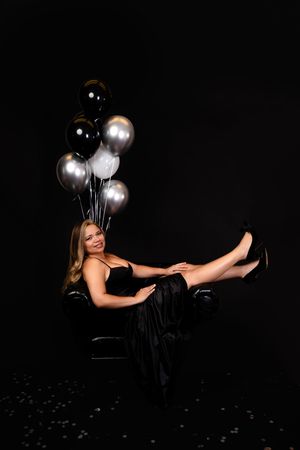 Woman in black gown sitting in chair with black, silver, and white balloons on dark background.