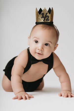 Adorable baby in black tuxedo bodysuit and gold glitter half birthday crown crawling on white background.