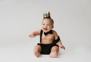 Adorable baby wearing a gold crown and black suspenders smiling joyfully against a white background.