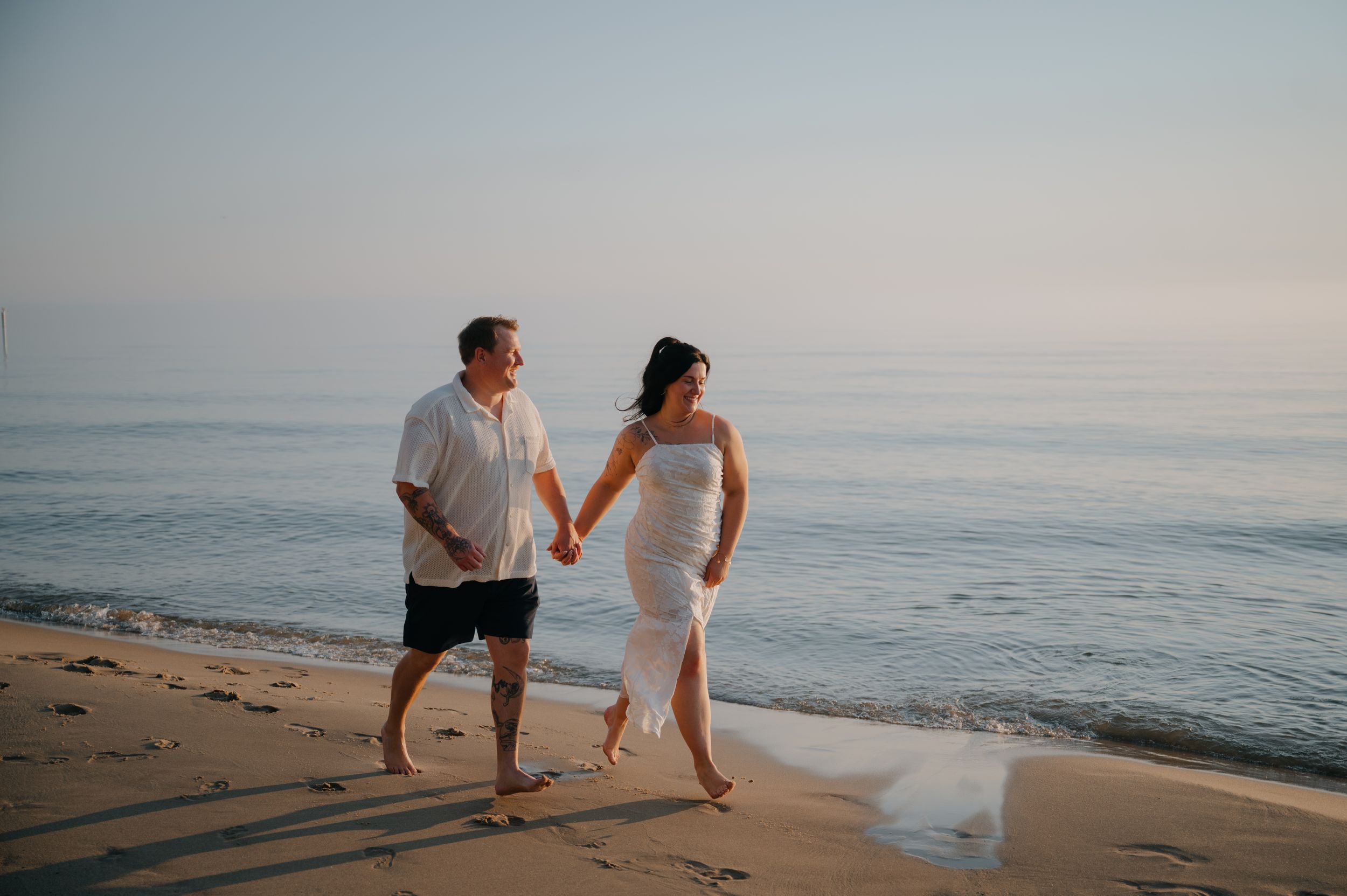 Couple in white outfits holding hands and walking barefoot along a calm sandy beach at sunset, with the bride's dress trailing behind.