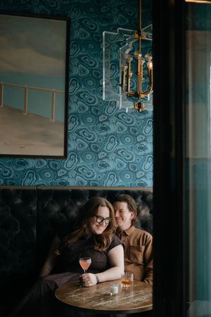 Couple sitting in a cozy tufted booth at a stylish bar with teal wallpaper, chandelier, and cocktails on a marble table.