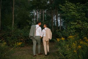Couple in neutral tones walks hand in hand along wooded trail with wildflowers.