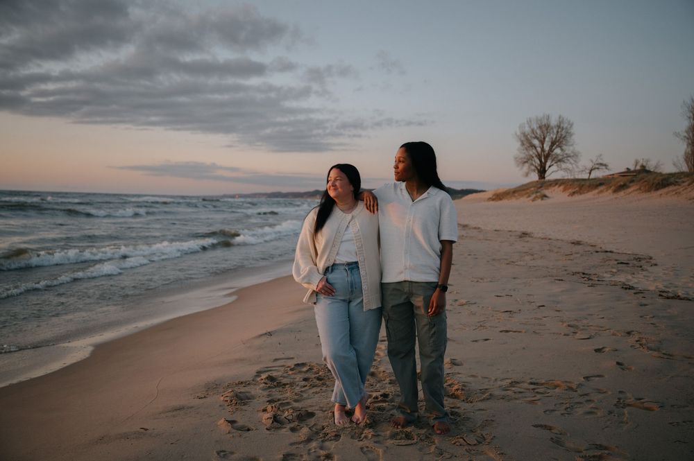 Couple standing together on a sandy beach at sunset, waves in background, wearing casual jeans and light tops.