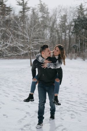 Couple sharing playful piggyback ride moment in snowy winter forest setting with evergreen trees in background.