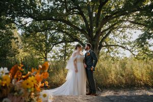 A couple in wedding attire stands together beneath a large tree with golden autumn foliage in the background.