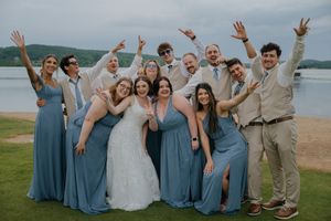Joyful wedding party in blue bridesmaid dresses and tan suits celebrates on a scenic beach overlook.