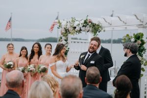 A waterfront wedding ceremony takes place with a bridal party gathered under a floral arch with boats visible in the harbor.