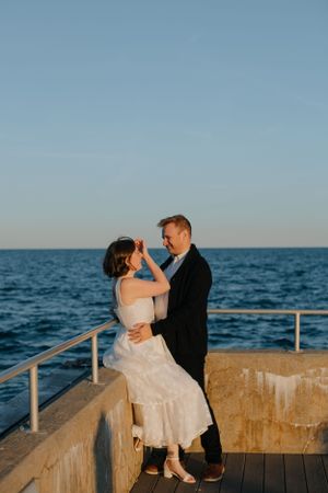 A romantic couple in wedding attire shares an intimate moment at sunset on an oceanfront pier.