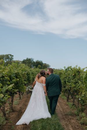 A couple in wedding attire walking through rows of grapevines at a vineyard on a sunny day.