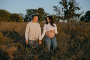 Expecting couple holds hands while walking through golden grass at sunset.