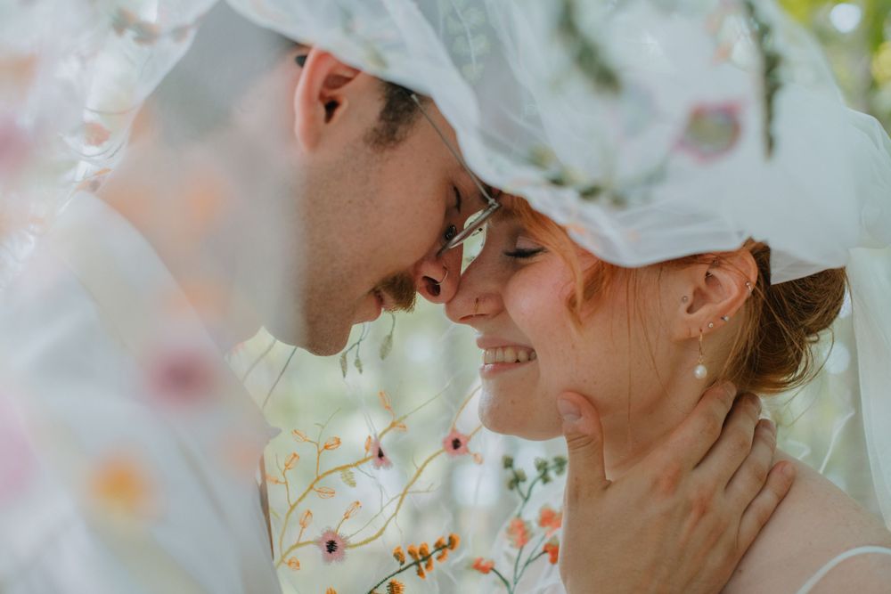 A romantic couple wearing white baseball caps share an intimate moment under a sheer veil with soft bokeh lighting.