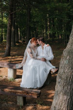 A romantic wedding couple sits intimately on a wooden bench in a sunlit forest setting.