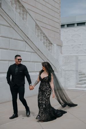 Couple in formal attire holding hands and walking against white stone building wall with long black veil flowing behind.