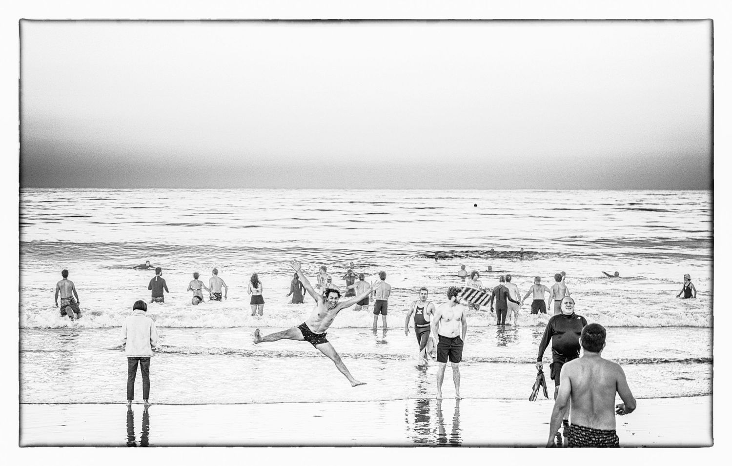 Black and white beach scene with people playing and swimming in the ocean waves.