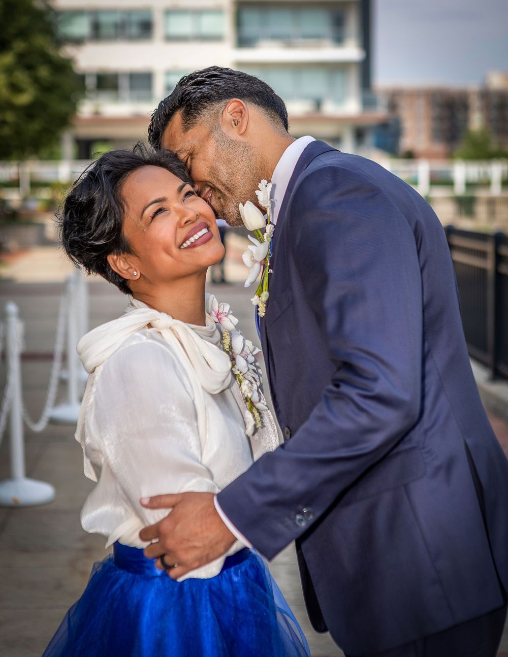 Couple embracing outdoors, bride in blue tulle skirt and white blouse, groom in navy suit with boutonniere.