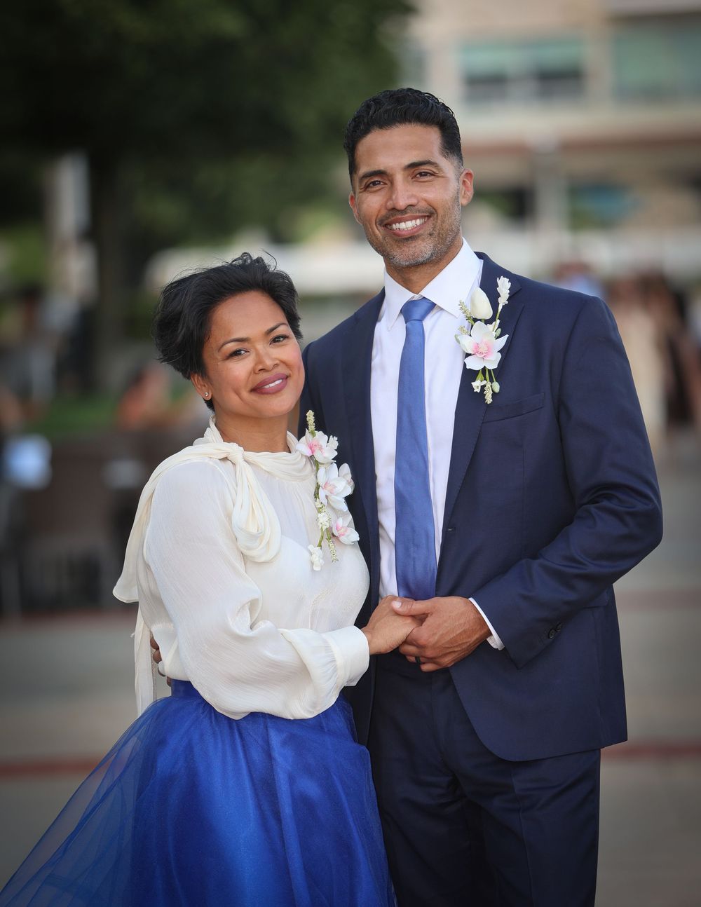 Elegant couple posing outdoors in formal attire, woman in blue skirt and white blouse, man in navy suit with boutonniere.