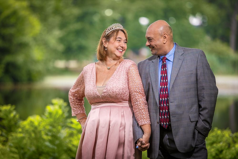 Couple holding hands outdoors, woman in pink dress with tiara, man in gray suit with red tie, green trees background.