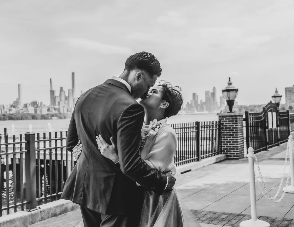 Black and white photo of a couple sharing a romantic kiss on a waterfront promenade with a city skyline backdrop.