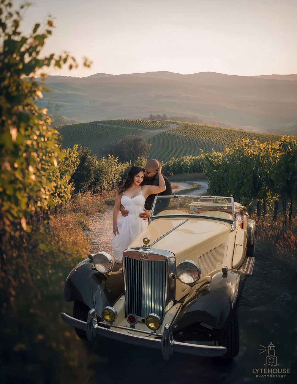 A couple poses with a vintage cream-colored luxury car against the backdrop of rolling hills at sunset.
