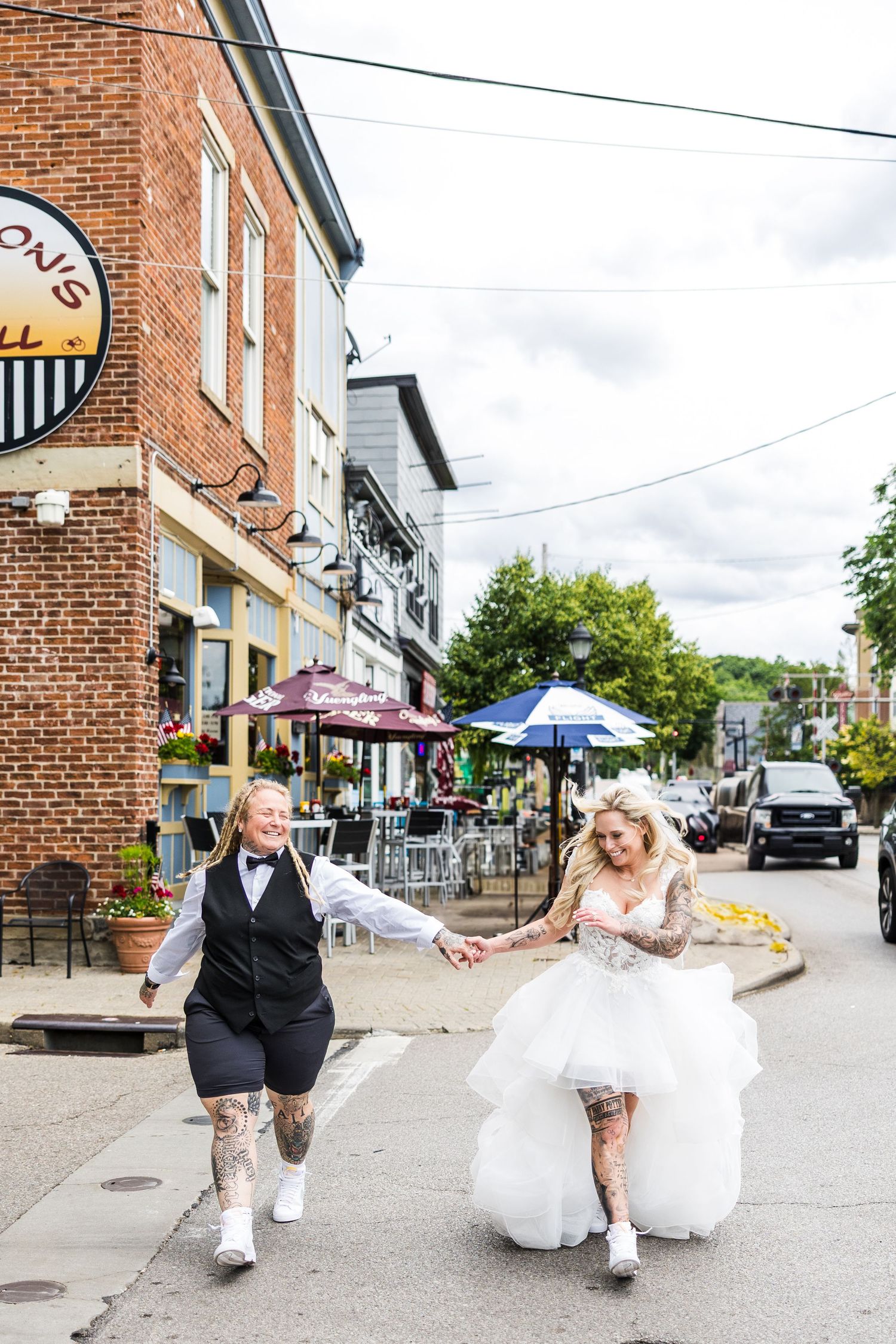 Two brides with tattoos run hand-in-hand down a small town street, one in a white gown and one in a black vest.