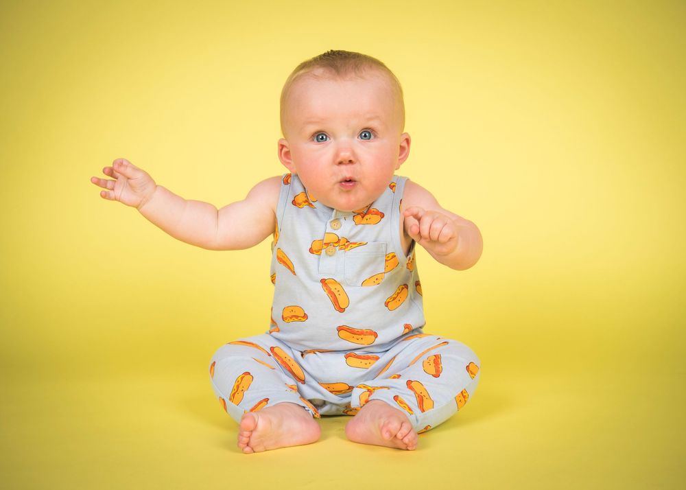 Baby wearing a food-patterned onesie sits against yellow background in progressive movement sequence.