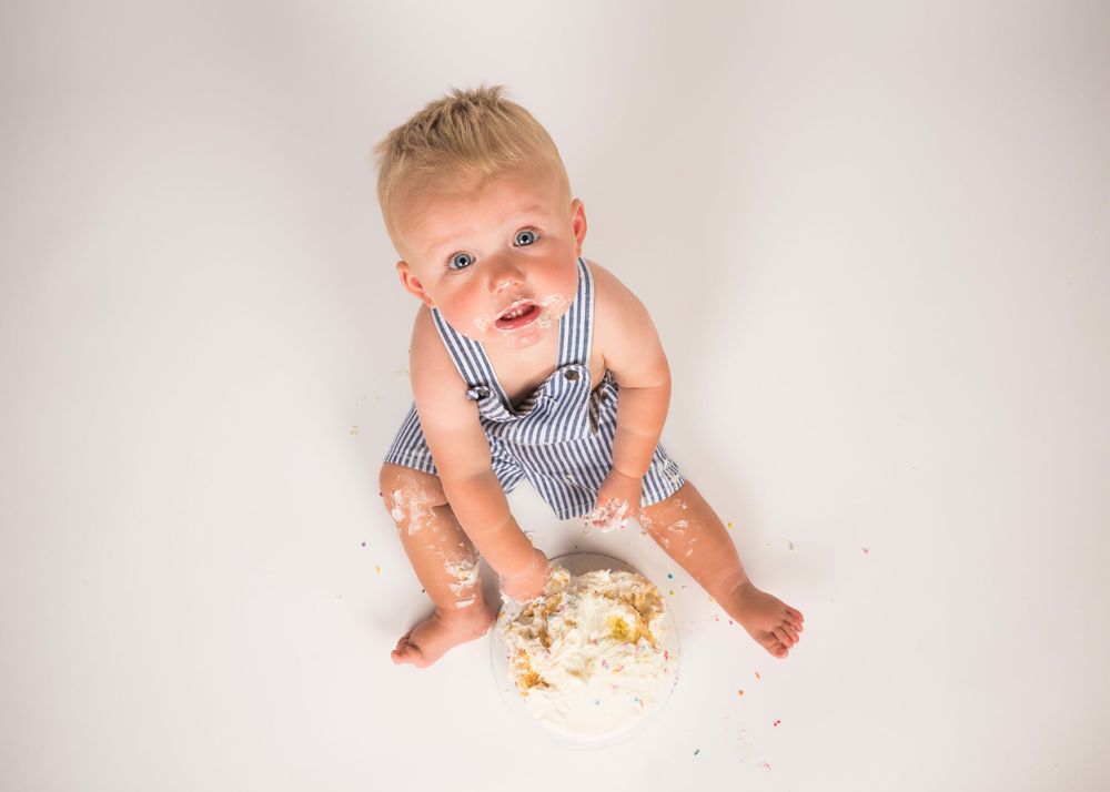 A baby in striped overalls sits on a white background next to a smashed cake during a cake smash photo session.