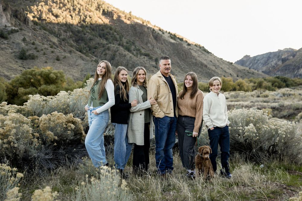 A group of people in casual attire pose together against a scenic mountain backdrop during golden hour.