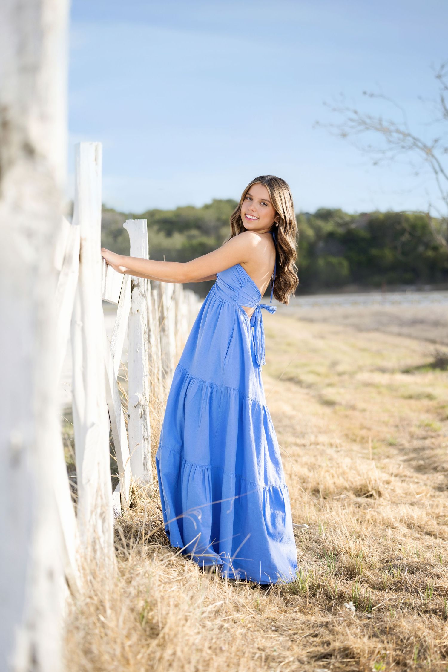 Young woman in blue maxi dress smiling by white fence in sunny field.