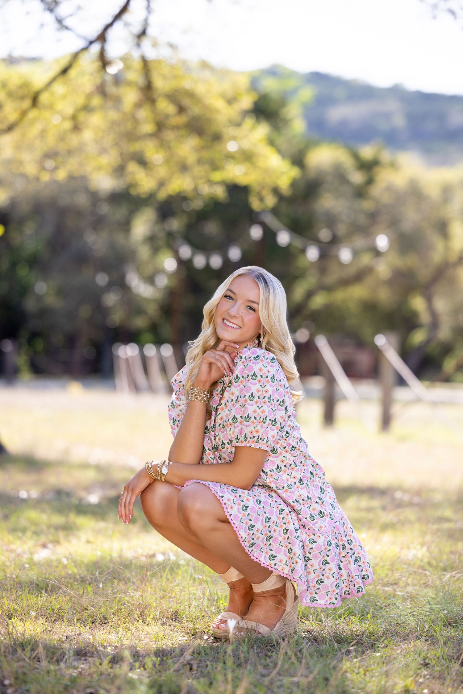Smiling blonde woman in floral dress crouching outdoors in sunlit field with trees and string lights.
