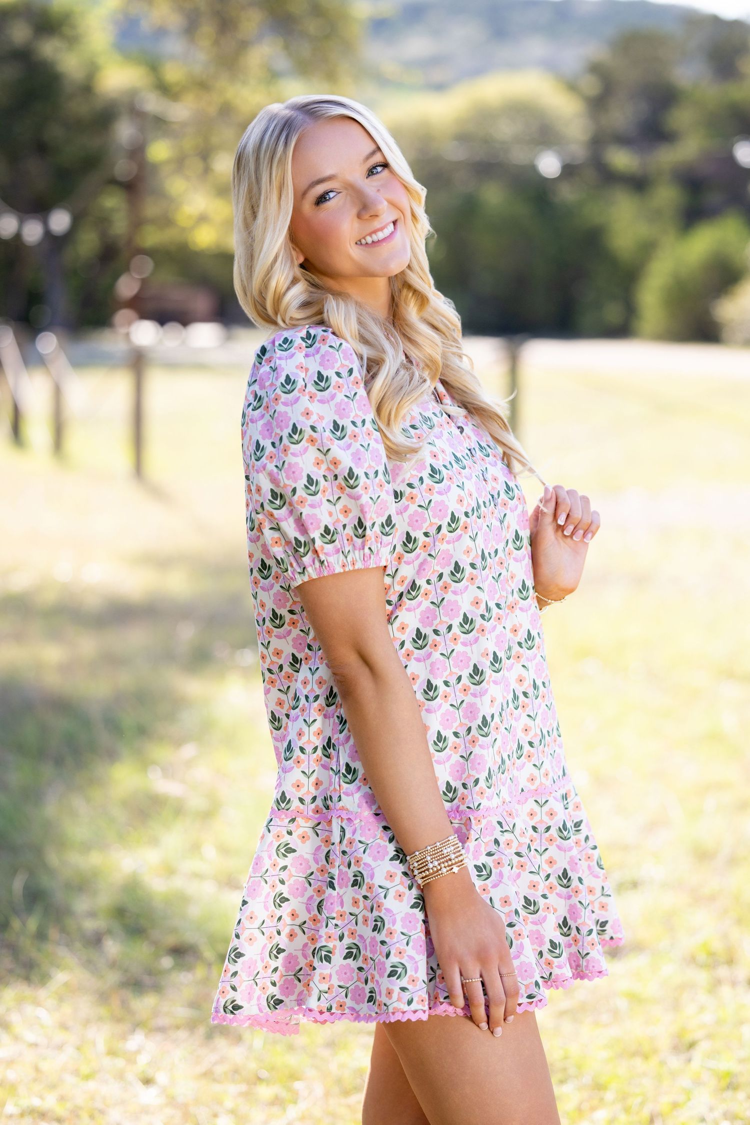 Blonde woman smiling in a pink floral mini dress, posing outdoors in a sunny green field.