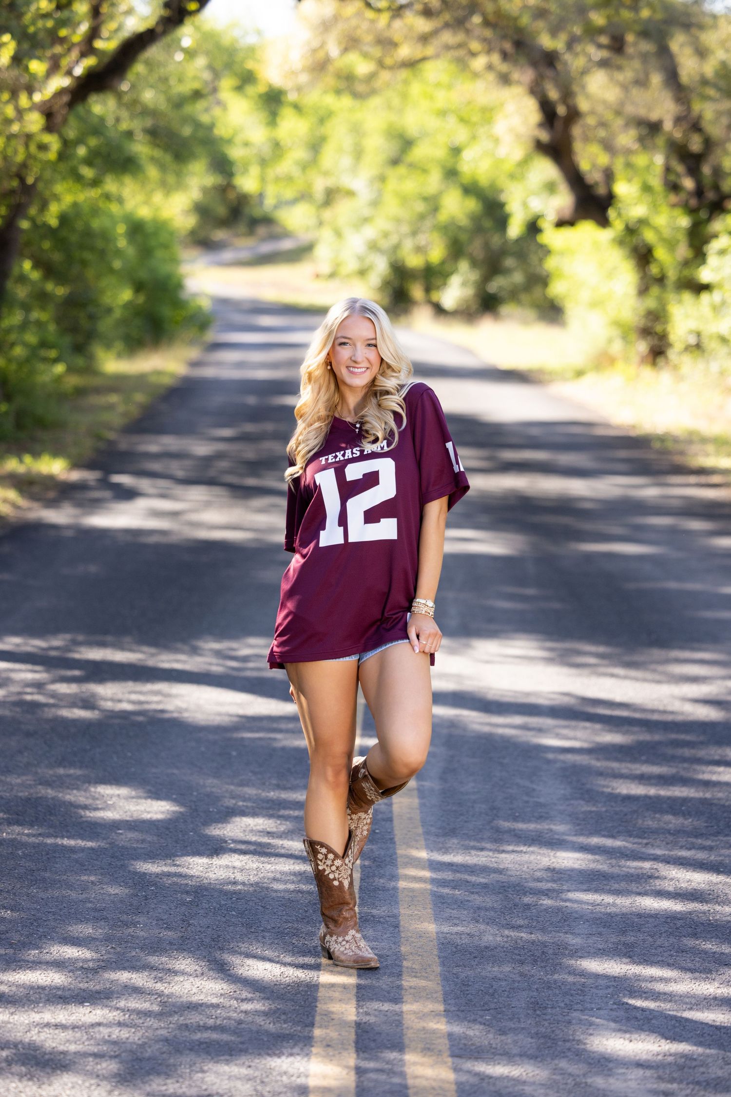 Blonde woman in Texas A&M jersey number 12 posing on a tree-lined road wearing cowboy boots.