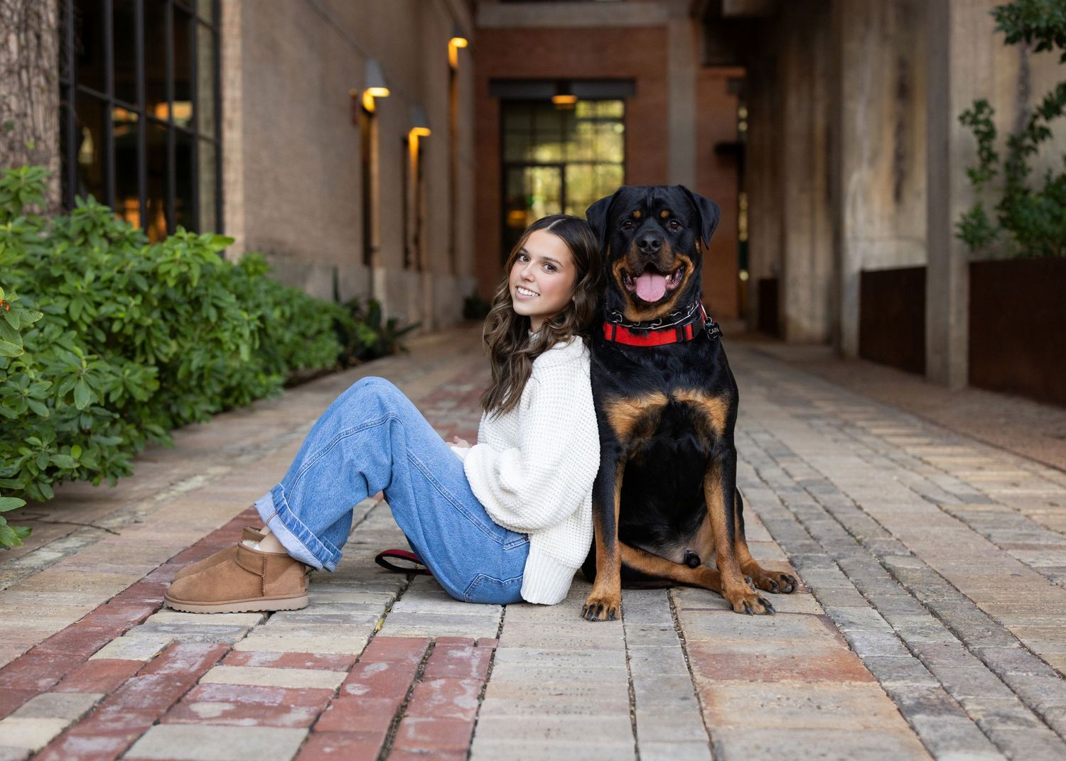 Young woman in white sweater and jeans sits smiling with a Rottweiler dog on a brick courtyard.