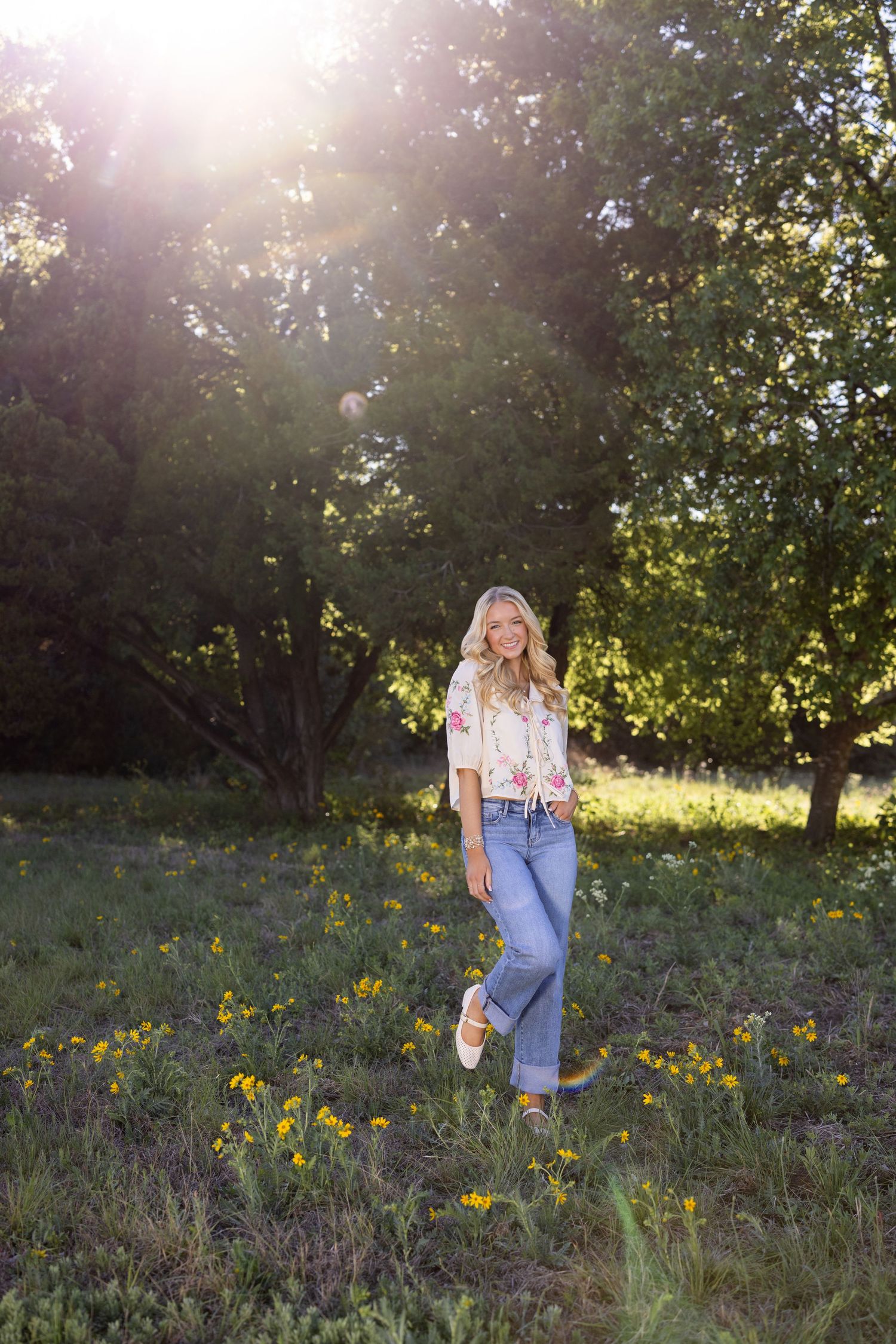 Blonde woman in floral top and wide-leg jeans standing in sunny meadow with wildflowers and green trees.