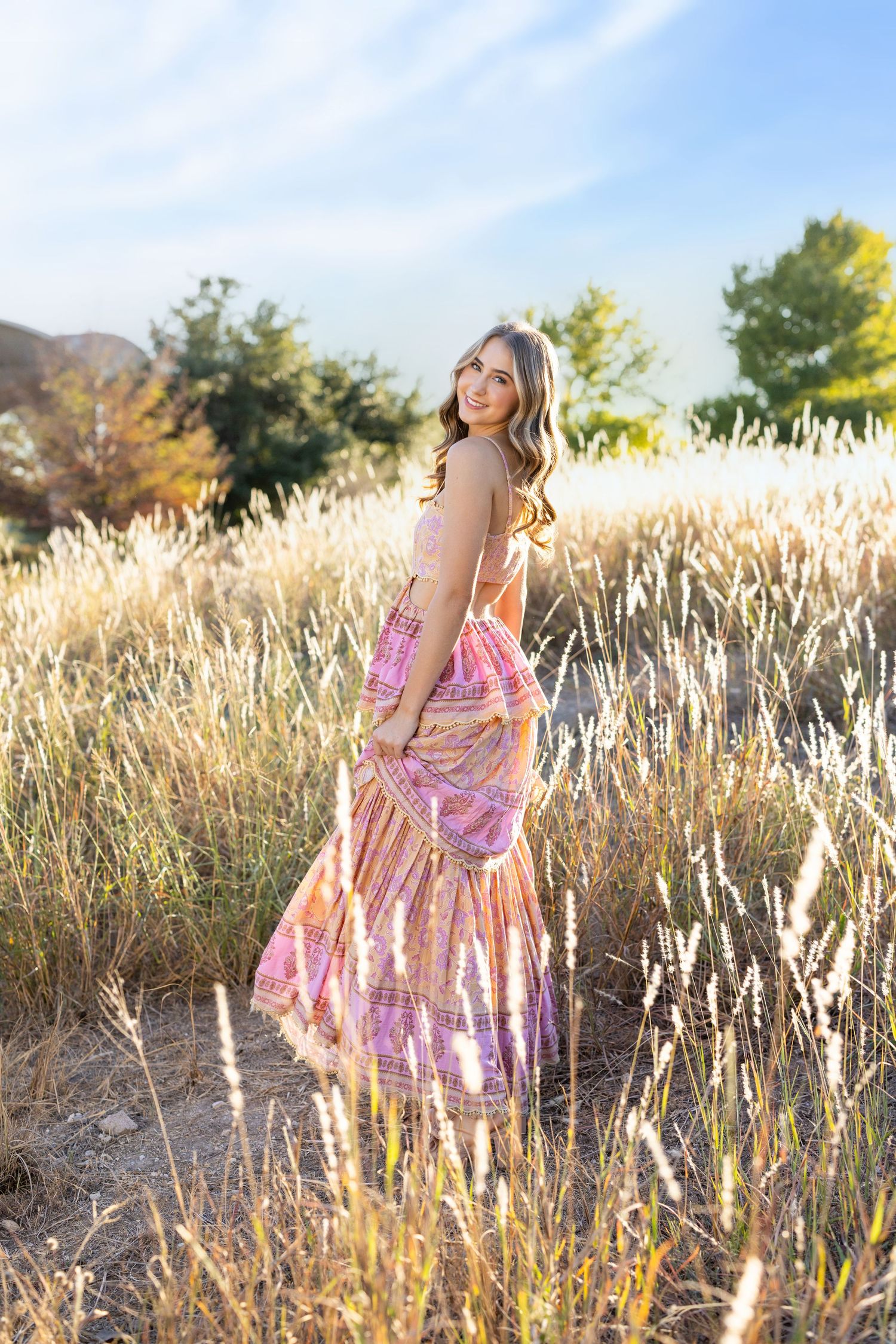 Young woman in pink boho maxi dress standing in golden field at sunset.