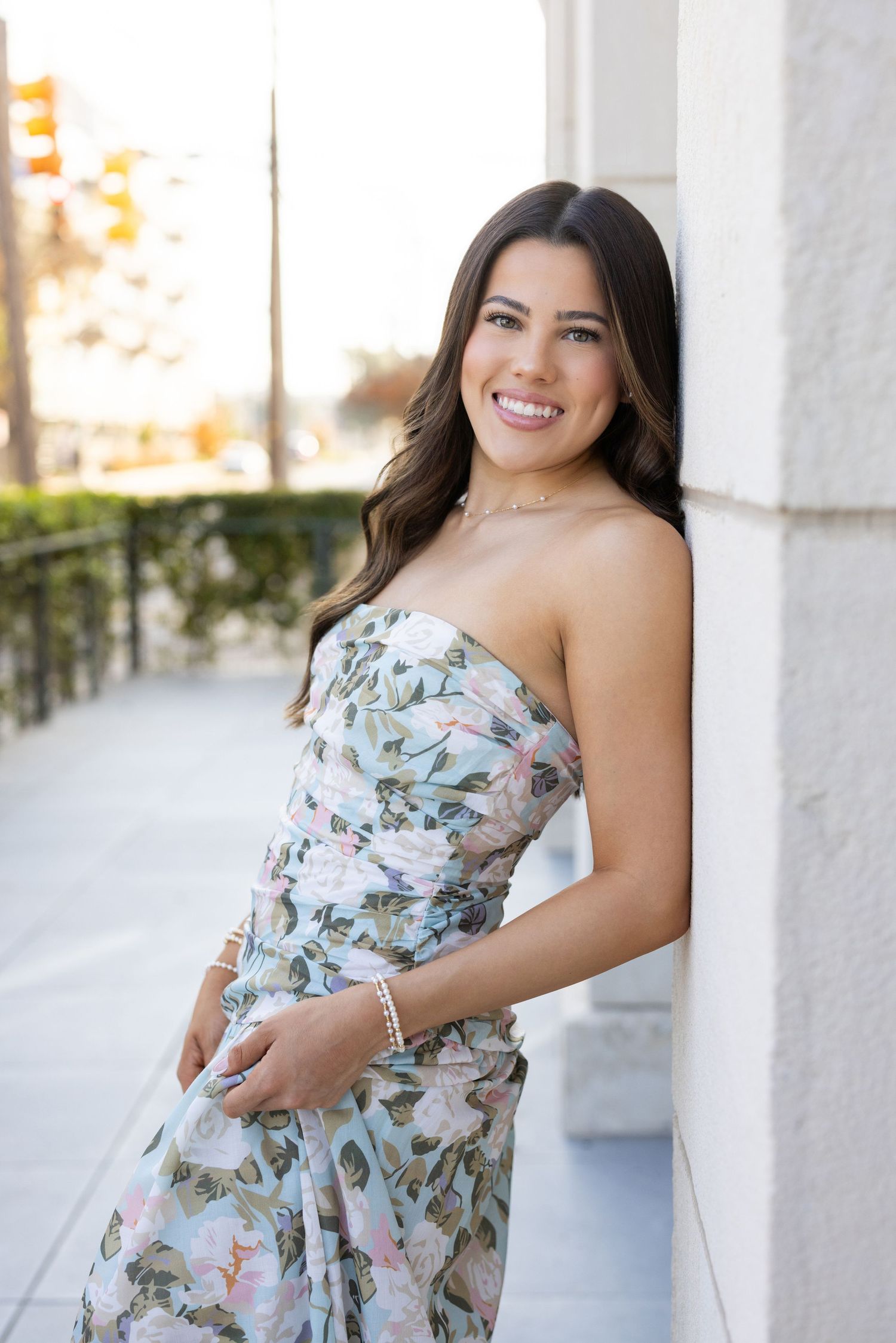 Smiling woman in strapless floral dress leaning against a white pillar on an urban street.