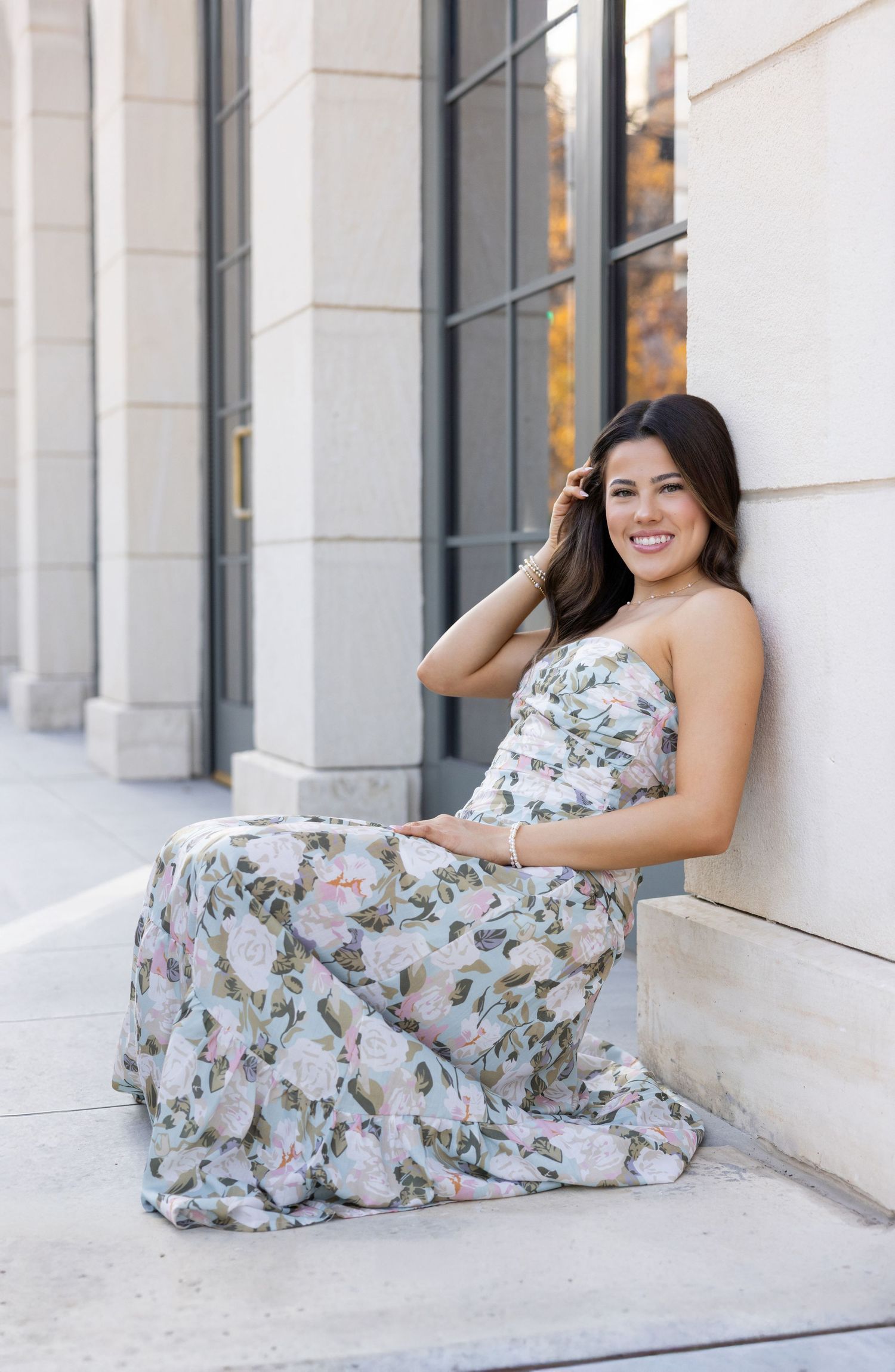 Woman in floral maxi dress sitting against a stone wall, smiling outdoors in natural light.