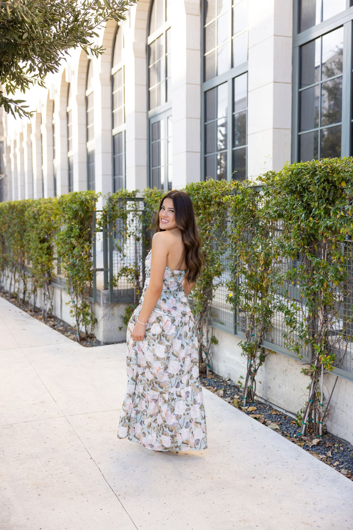 Woman in floral maxi dress posing on sidewalk beside ivy-covered stone building, looking back over shoulder.